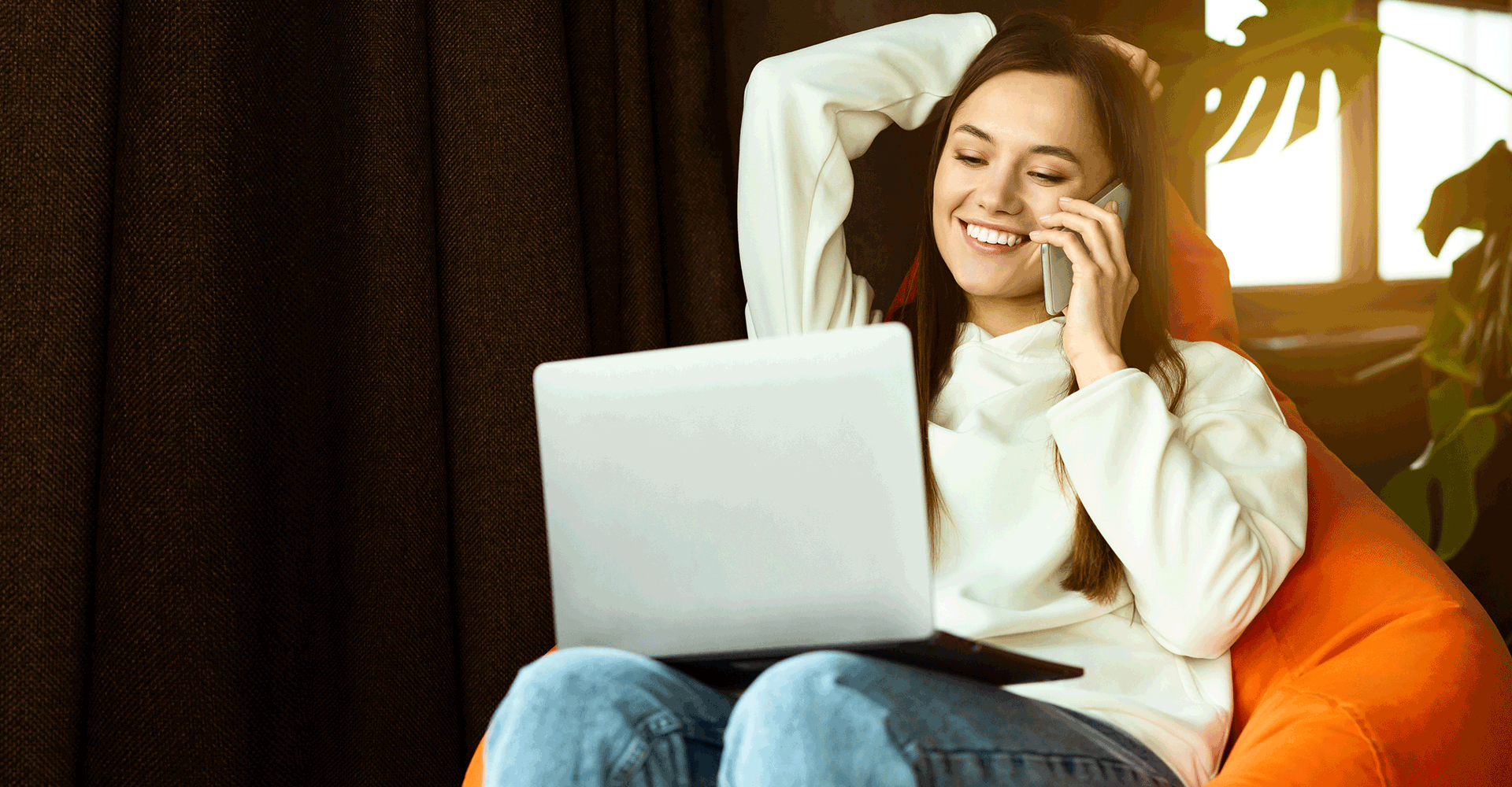 Nainen käyttää hymyillen tietokonetta ja puhuu puhelimessa.